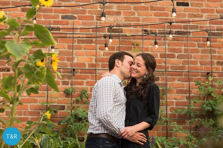 A man kisses the cheek of his fiance in front of a red brick building in Corktown