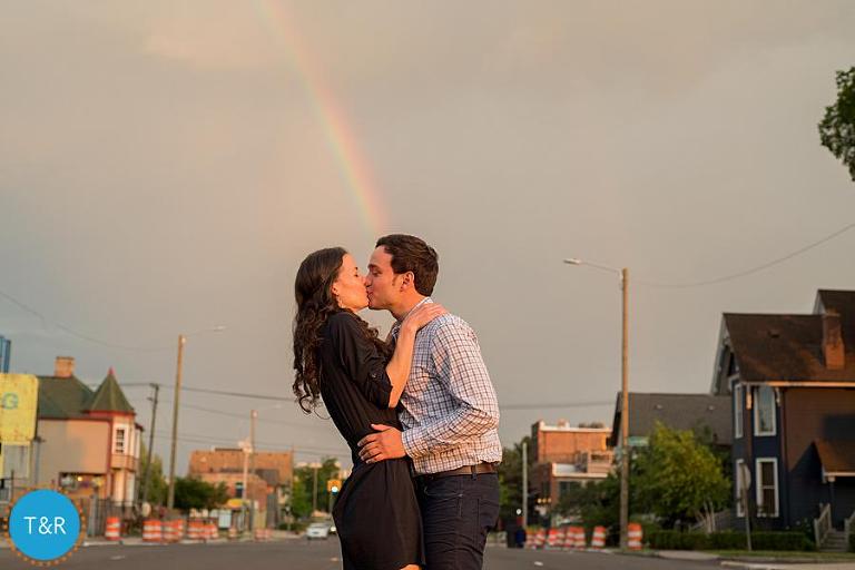 A couple kisses on the street with a rainbow behind them 