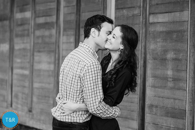 A man kisses his fiancé on the cheek in front of a wooden wall